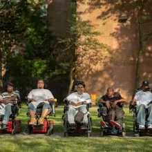 Five Black and brown men (from left to right: Vince, Jay, Tito, Pete and Var) sit in wheelchairs outside, lined up in a straight horizontal line facing the camera. They wear T-shirts and sneakers and some wear baseball caps. They all look into the camera with serious expressions. 