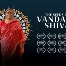 Vandana Shiva wears a red sari and stands in front of a halo of seeds. 