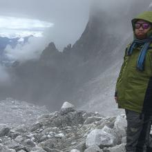 Phuntsho Tshering, a man of South Asian descent, is standing on top of a rocky terrain in the rugged mountainous Himalayas looking straight into the camera. He is wearing shades, a pair of gumboots and hiking clothes. 