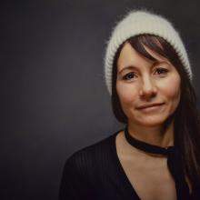 Headshot of a Latina woman with olive skin an long brown hair wearing all black and an off-white cashmere beanie. 