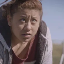 A Latina teenager leans over in a field, tilting her head upwards and looking away from her harvesting work.