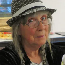 An older White woman in glasses and hat reads from a composition book before a notebook.