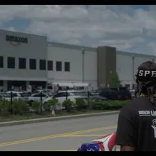 A man wearing an "Amazon Labor Union" t-shirt looking towards an Amazon Fulfillment Center warehouse. 