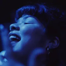 Latina woman dances to music amongst a crowded concert audience, in blue light. 