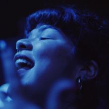 Latina woman dances to music amongst a crowded concert audience, in blue light. 