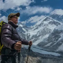 Renowned climber Minga Tsiri Sherpa stands holding walking poles in his hands and wearing a backpack as he looks out towards the Himalayan mountains which are also seen behind him in the distance. 