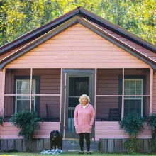 An older woman in a pink cardigan stands in front of a pink house with her dog.
