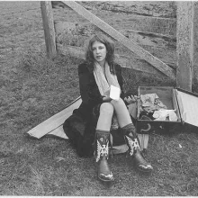 Young woman in early 70's fashion sits on a picnic blanket next to an open suitcase with a notepad in her hand.