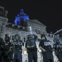 A line of Baltimore police officers wearing riot gear and holding shields stand in front of city hall the night after the murder of George Floyd.