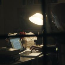 A woman in a hijab working at her computer late into the night, slightly obscured by a windowpane in the foreground. 