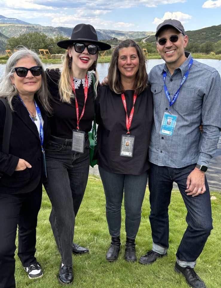 On the grassy banks of a lake, four white people wearing festival lanyards stand casually and happily.