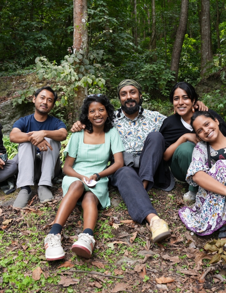 Six brown-skinned individuals (three men, three women) sit on a grassy knoll in a forest, smiling at the camera