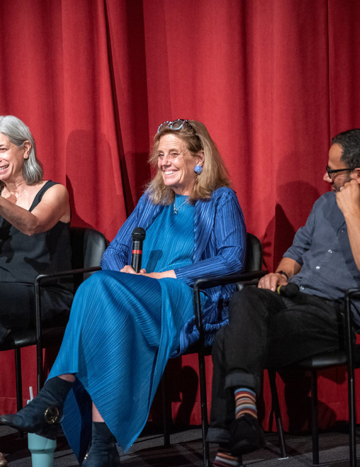 In front of red curtains, three people sit on chairs on a stage.