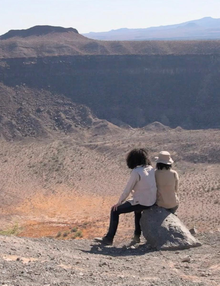 Two people sit on a rock against a vast desert landscape before them
