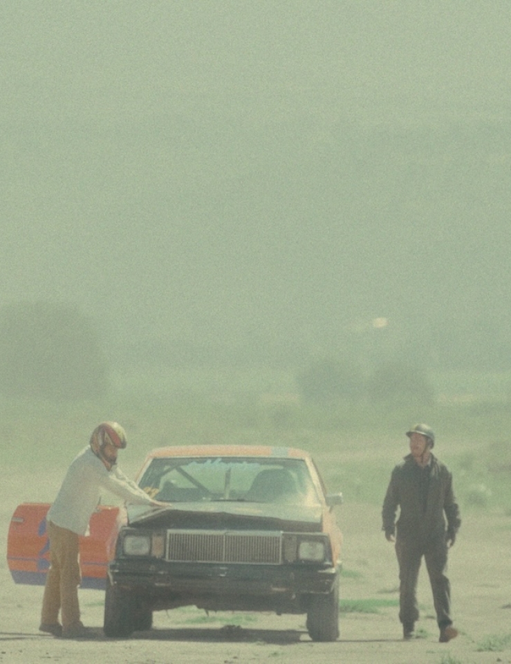 In a dusty road, two men with helmets on stand on either side of a broken down, battered car.