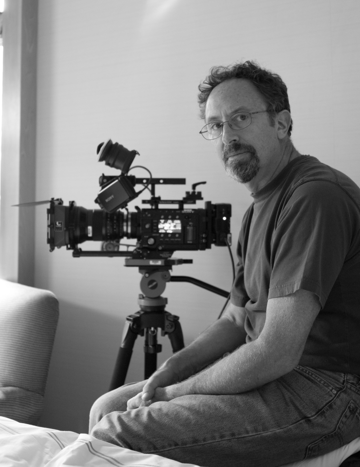 Black and white image of a white man with short hair, glasses and goatee, man sitting in a room next to a camera, as another stands by a window.