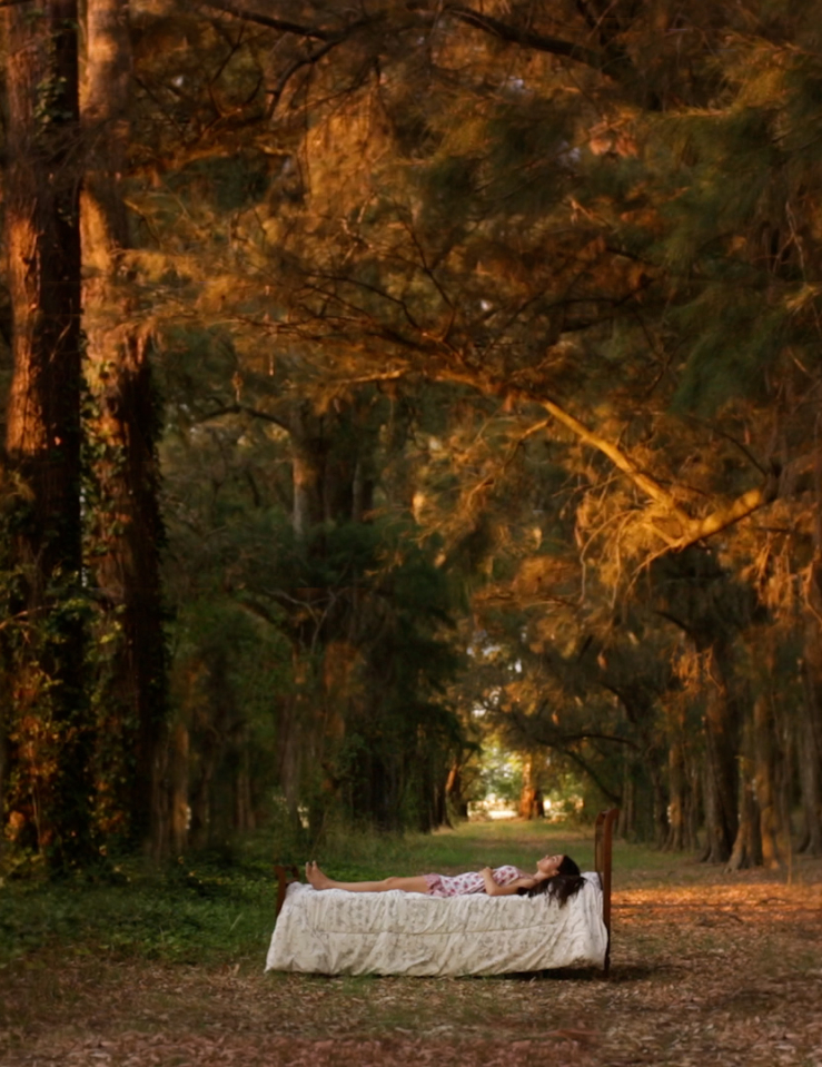 A woman lays on a bed that stands alone in the middle of a dusk-lit forest
