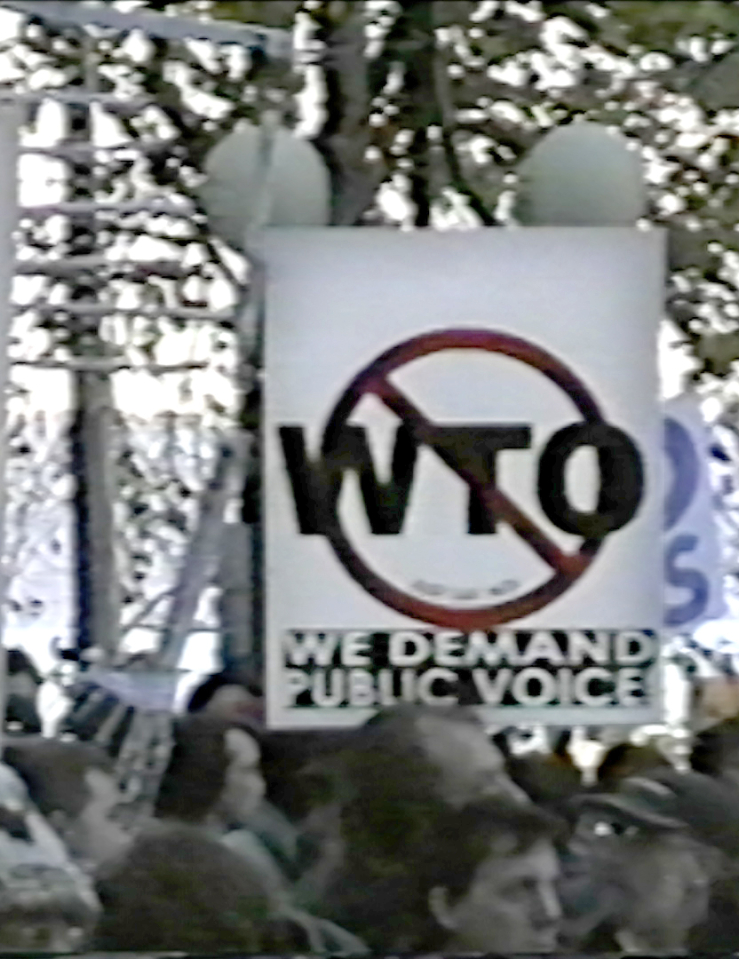 Grainy shot of group of outdoor protestors holding "We Demand Public Voice" signs with WTO crossed out