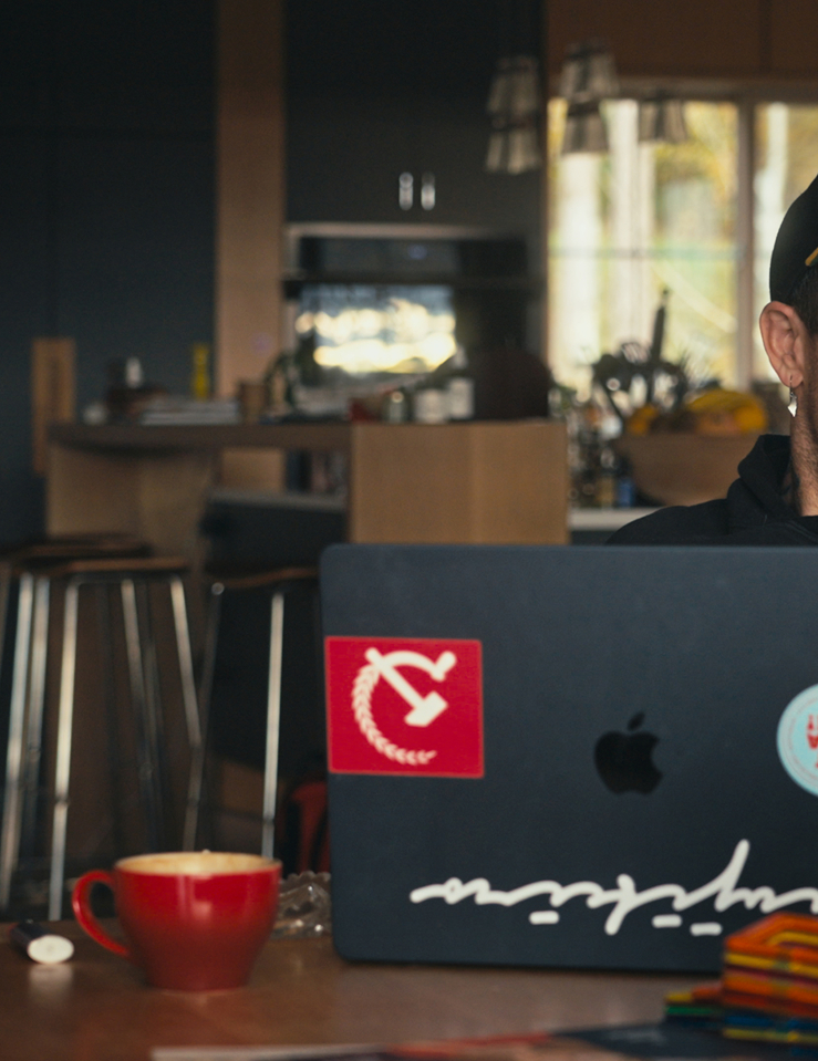 A white man wearing a cap and a hoodie sits at a kitchen table with his laptop, which is adorned by a communism sticker and another that reads "Let Cuba Live"
