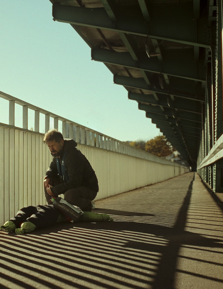 Wide shot of a bearded middle-aged white man with short-cropped hair kneeling over a mannequin of sorts at the railing on a bridge
