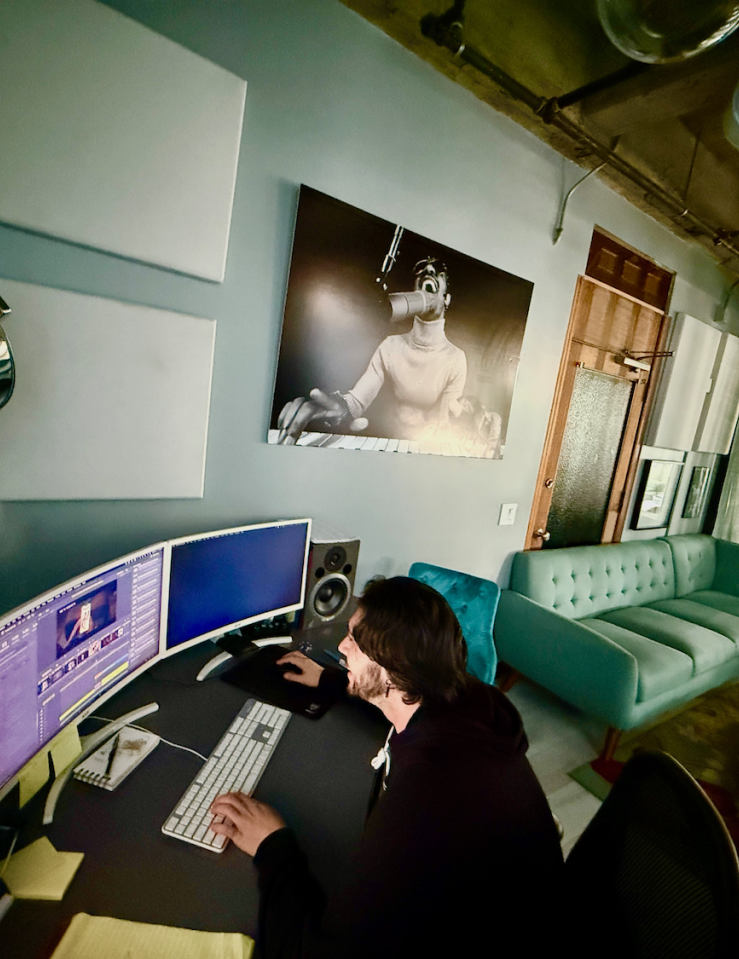 A white man with long black hair in a hoodie sits at an editing bay in a stylish, industrial-looking, emerald-decorated room