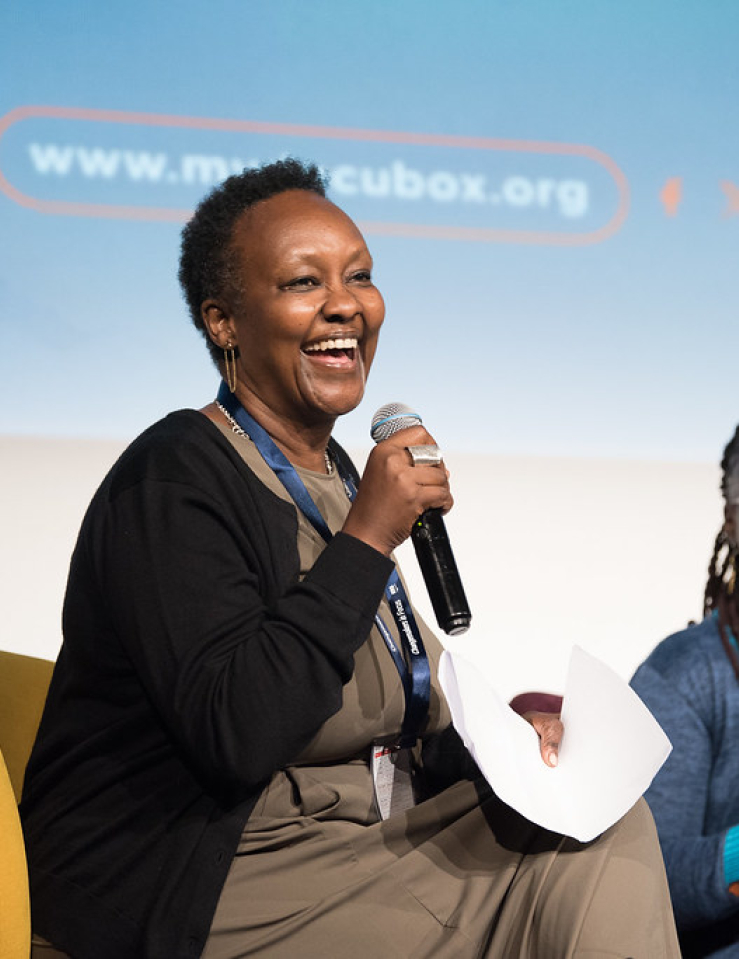 A Black woman with short-cropped hair sits at a stage next to another Black woman; both are holding mics and smiling widely