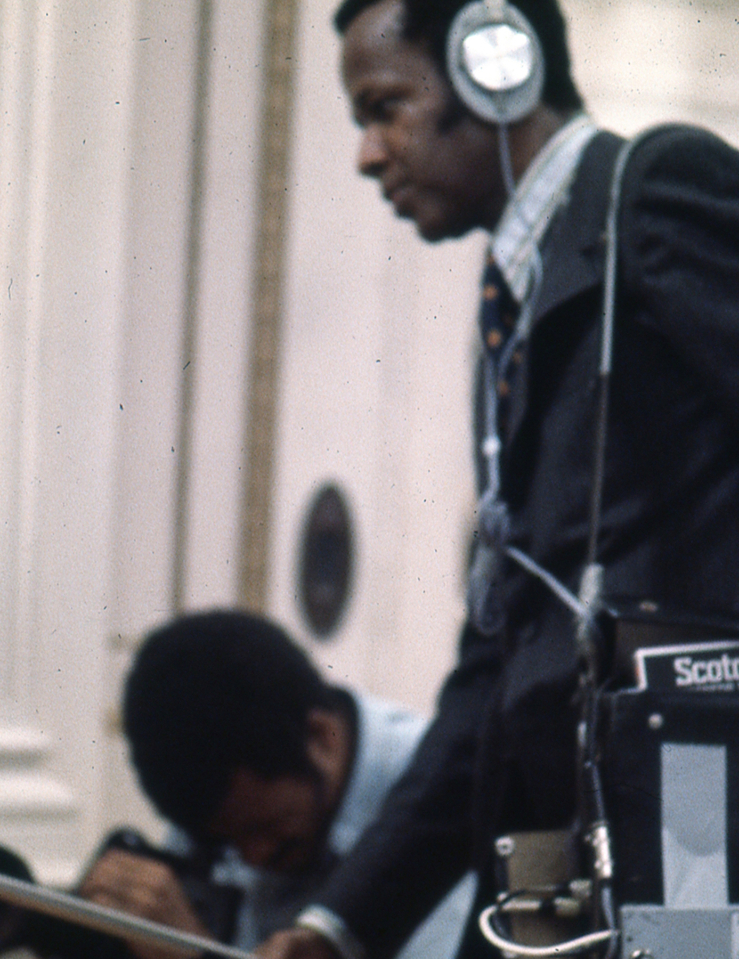 One man looks through a film camera viewfinder while another holds a microphone and sound recorder while wearing headphones.