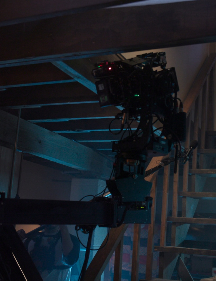 A young white actor wearing a blue button down shirt and pants stands on a basement with a camera rigged in front of him