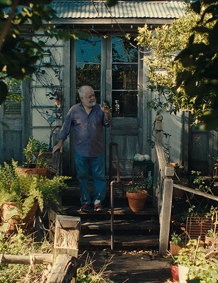 A man in blue jeans and a long sleaved shirt stands in front of the door of his house and his lush, green front yard.