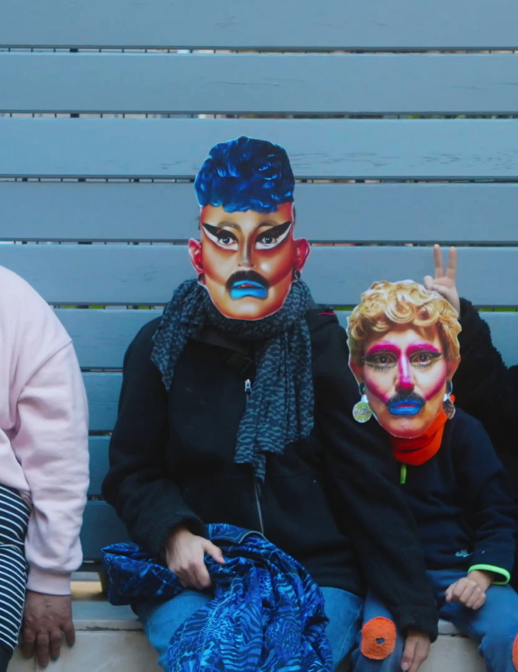 Four people sit against a light blue wall wearing regular clothes and paper masks of drag queens