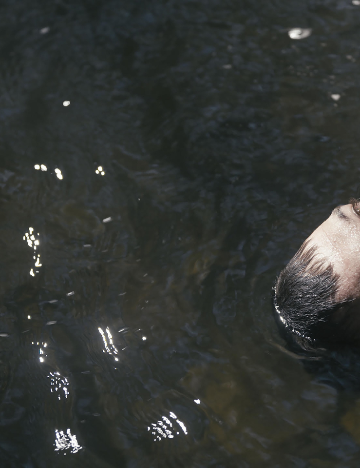 A white man with dark hair and hazel eyes looks up as he floats on a body of water