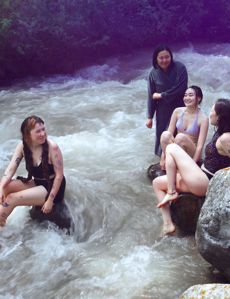 A group of soaked women gather around a riverbed, with pink smoke gathering around them
