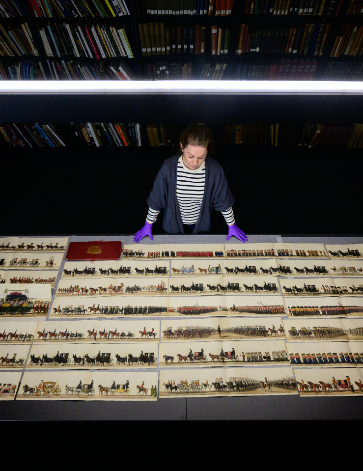 A white female archivist stands in front of a number of old illustrations of an 18th century English coronation, stacks like in a library behind her