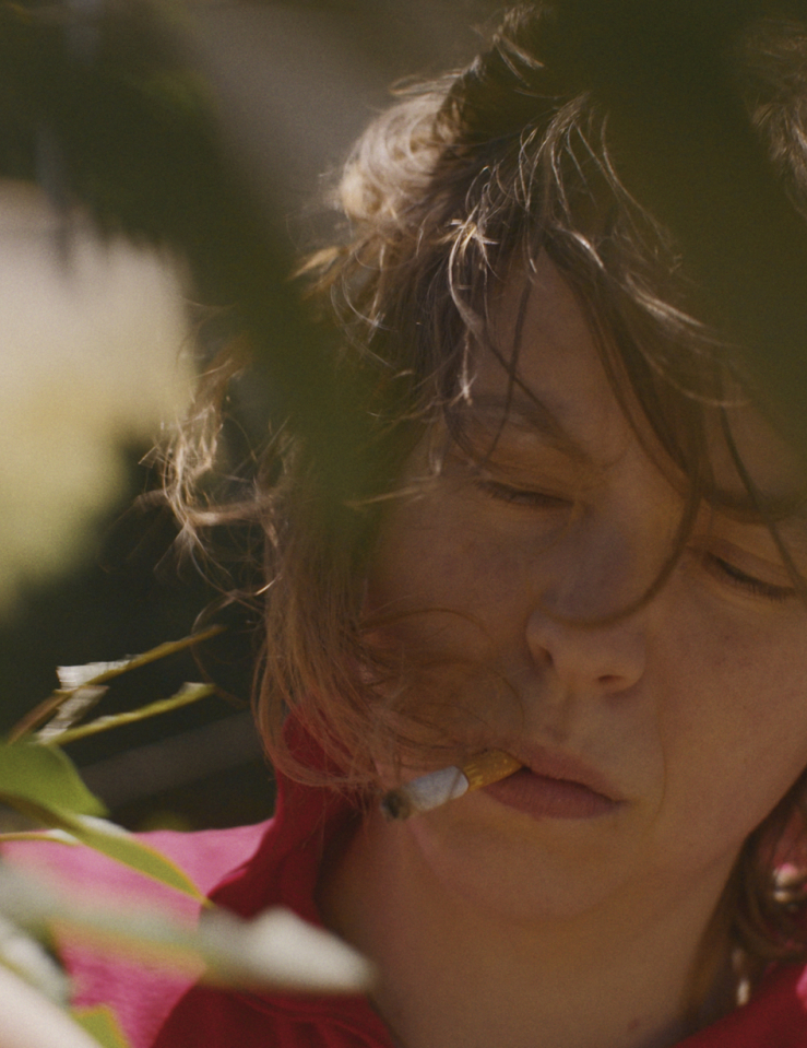 A close up of a white woman with shoulder-length dark hair in a red shirt, eyes closed, holding a cigarette in her mouth outside