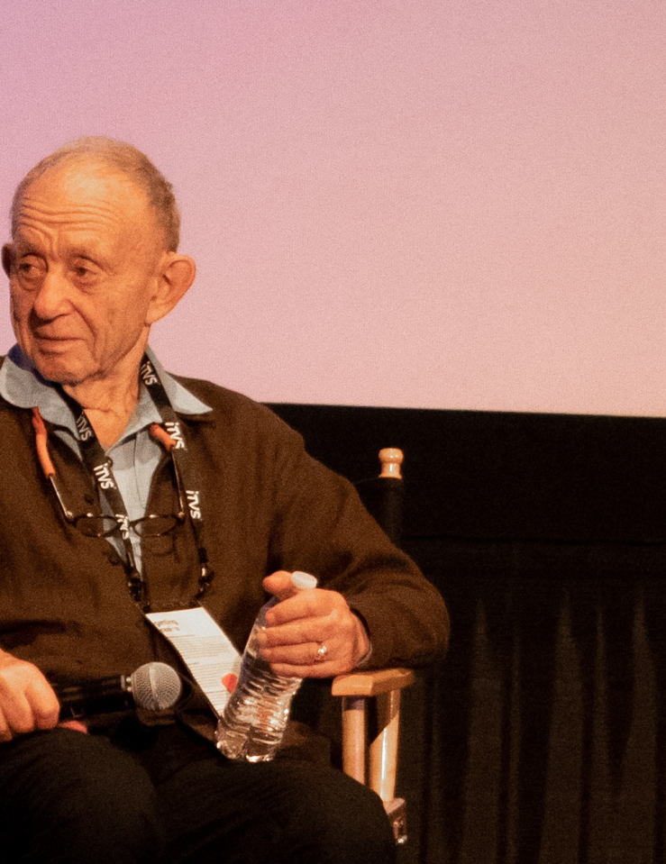 An older, balding white man with short wispy white hair wears a blue shirt and a brown cardigan while sitting on a director's chair on a stage, holding a mic and a water bottle.