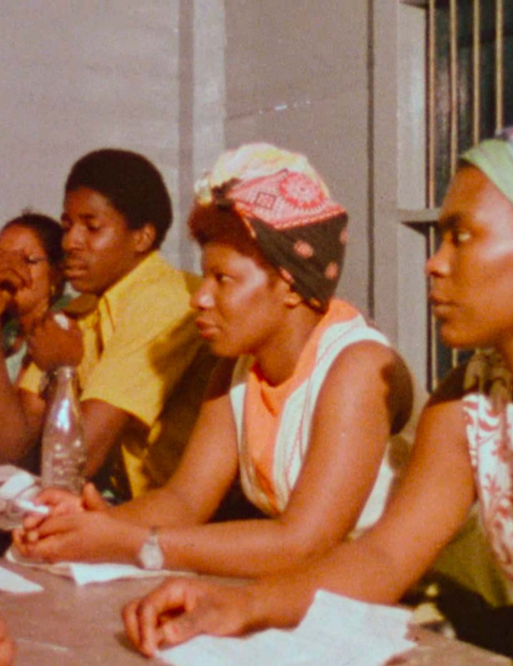 A group of Black women (in bright colorful sleeveless dresses and headwraps) sit at a table, as if in a meeting