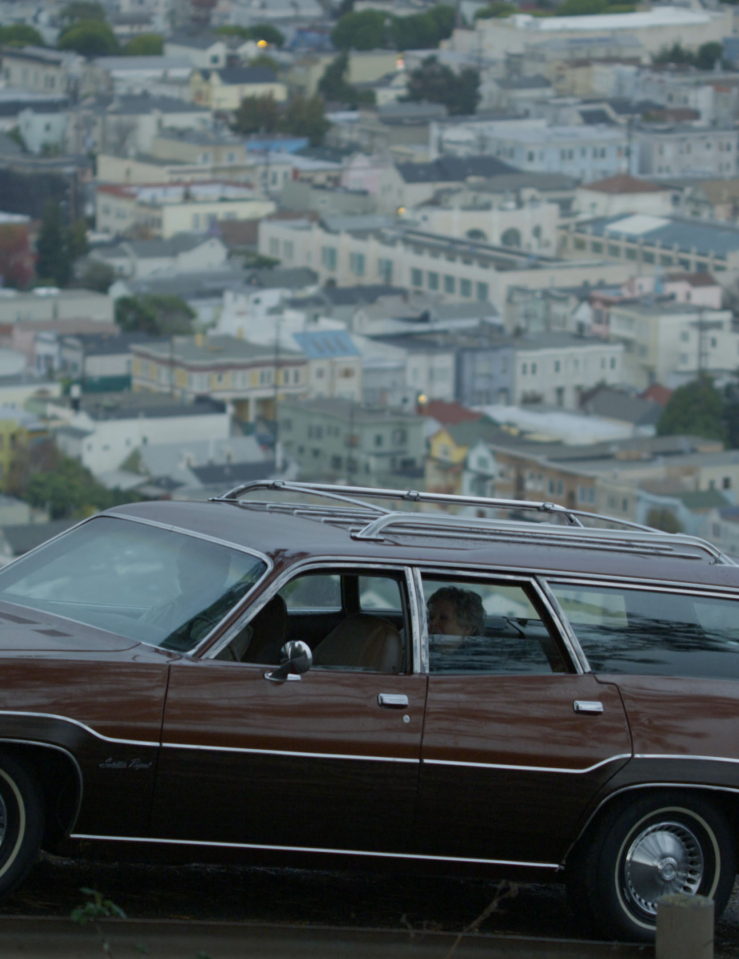 A brown station wagon is parked atop a street that overlooks a city down below; an older woman can be seen sitting in the backseat while a man stands nearby in the foreground