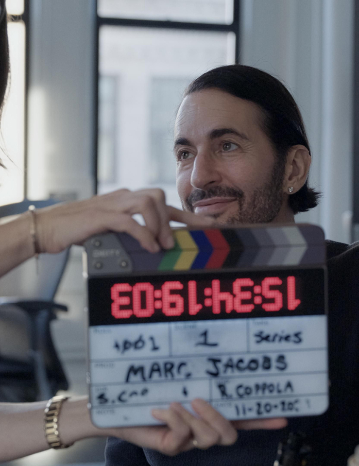 A middle-aged white man with slicked back black hair and five o'clock shadow sits in front of a camera as a white woman with shoulder-length brown hair holds a clacker that reads "Marc Jacobs" in front of him