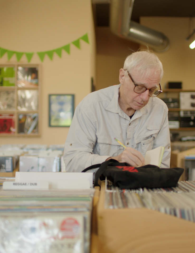 An elderly white man with thin white hair and glasses jots something on a notebook while in the middle of an old-school record store