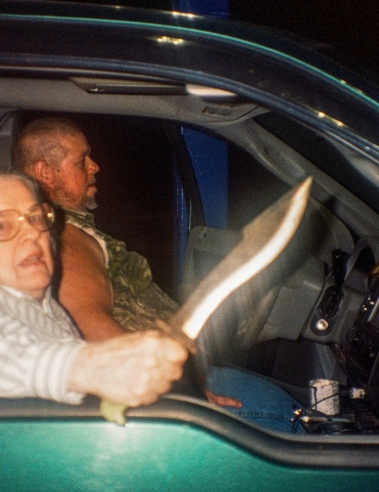 An older women with glasses sitting in a truck at night wields a knife at the camera
