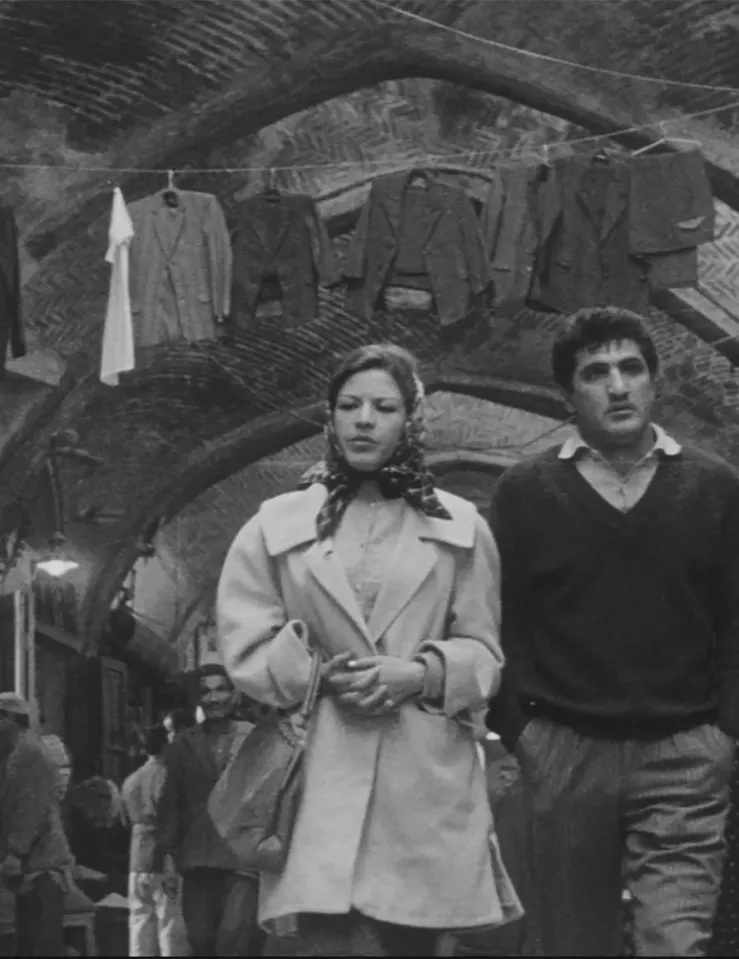 Black and white image of a woman in a coat and silk scarf over her hair walking alongside a man in pants ad a v-neck sweater around a Middle-Eastern market
