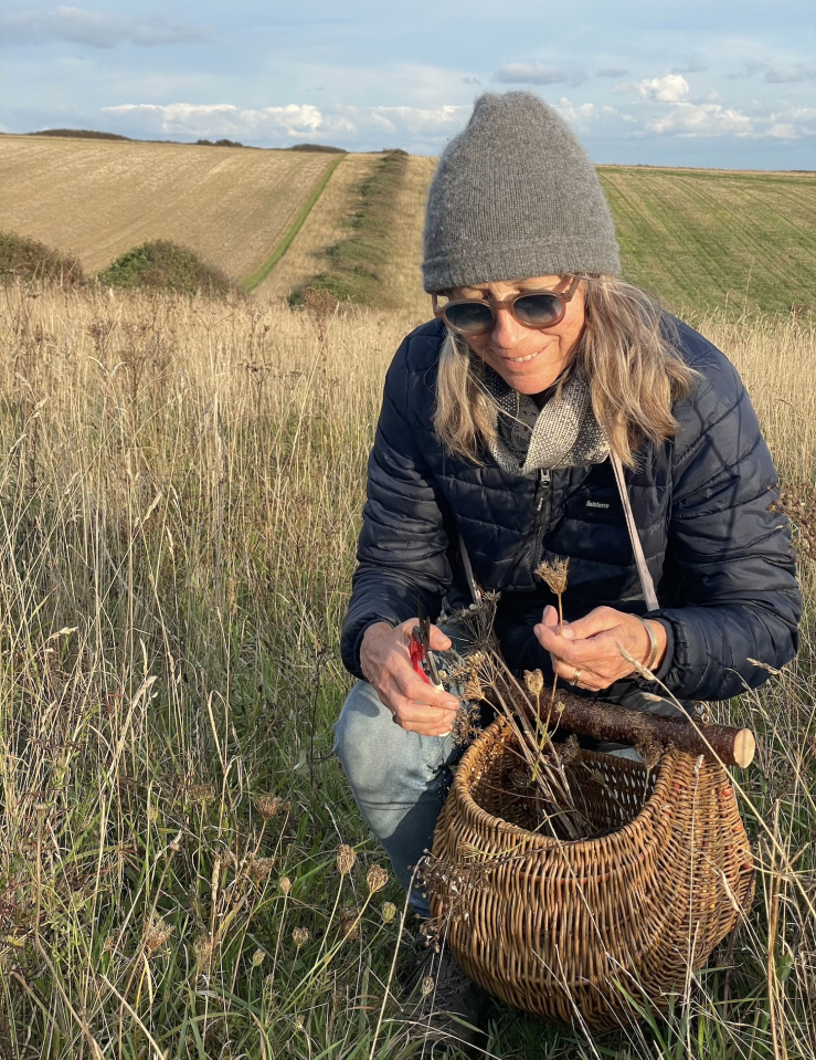 A middle-aged white woman with shoulder-length auburn hair wearing sunglasses, a winter hat and a warm jacket squats down as she gathers wild flowers in a field.