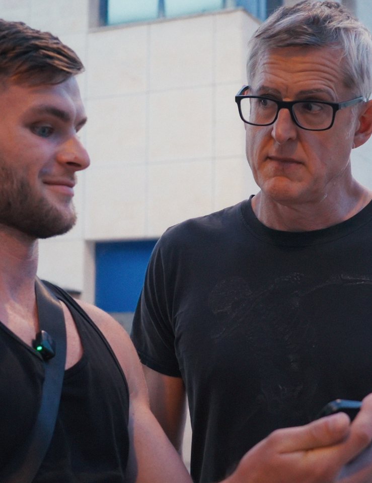 A middle-aged white man in a black t-shirt and rimmed glasses stares incredulously at a young muscled white man in a black tank top as he stares into his smartphone