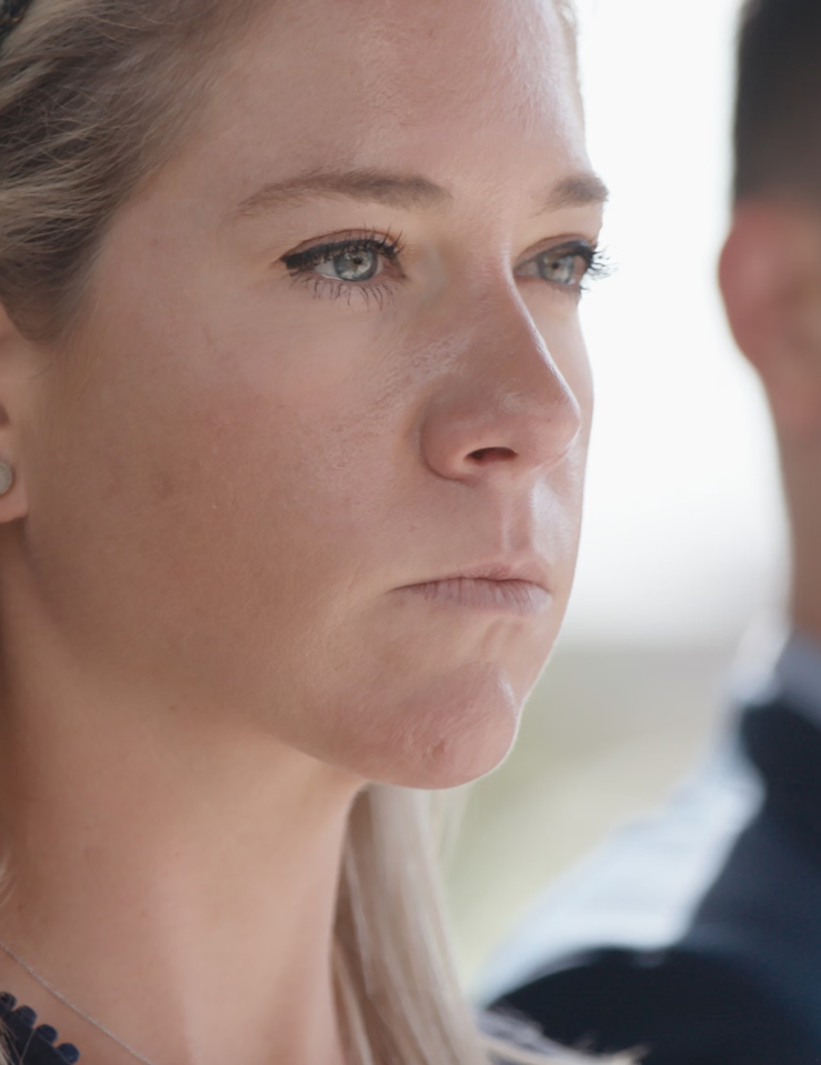 A young white woman with shoulder-length blond hair stares intently away from her; a young man in a suit is out of focus in the background