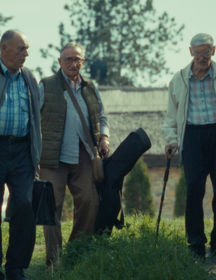 A group of elderly men in casual clothing carrying instruments in cases walk alongside a countryside road