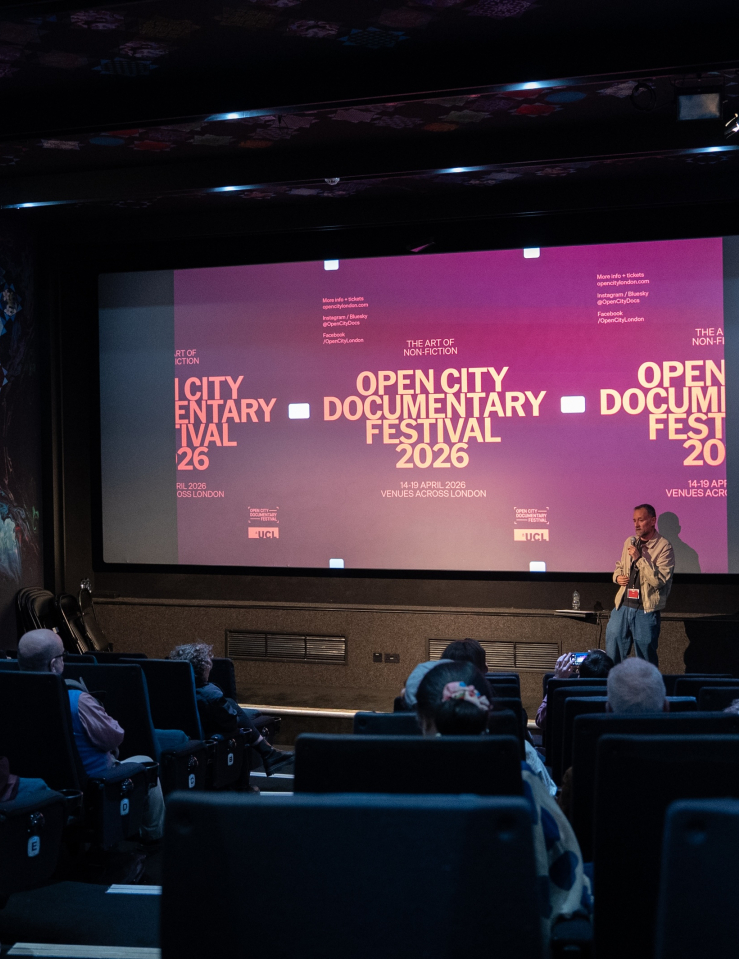 A white man stands in front of a crowded theater, a screen behind him showcasing an ad for the Open City Documentary Festival