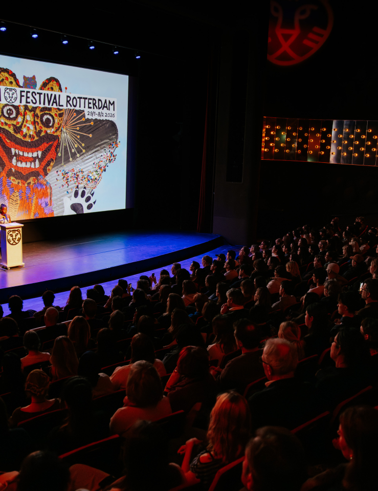 A young Black woman stands at a podium on a stage dwarfed by the audience in attendance with a sign for the International Rotterdam Film Festival behind her