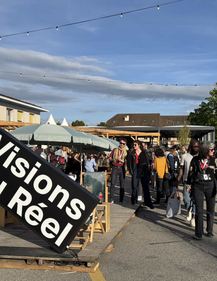 Under a crisp blue sky, a crowd of festivalgoers stands under the outdoor umbrellas. In front of them is a large sign that reads: "Visions du Reel."