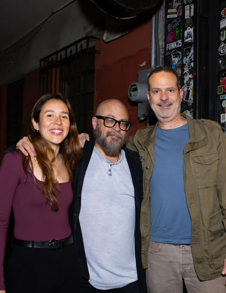 A young woman with long brown hair in a burgundy sweater poses arm in arm with two middle-aged men dressed casually in front of a local cinema