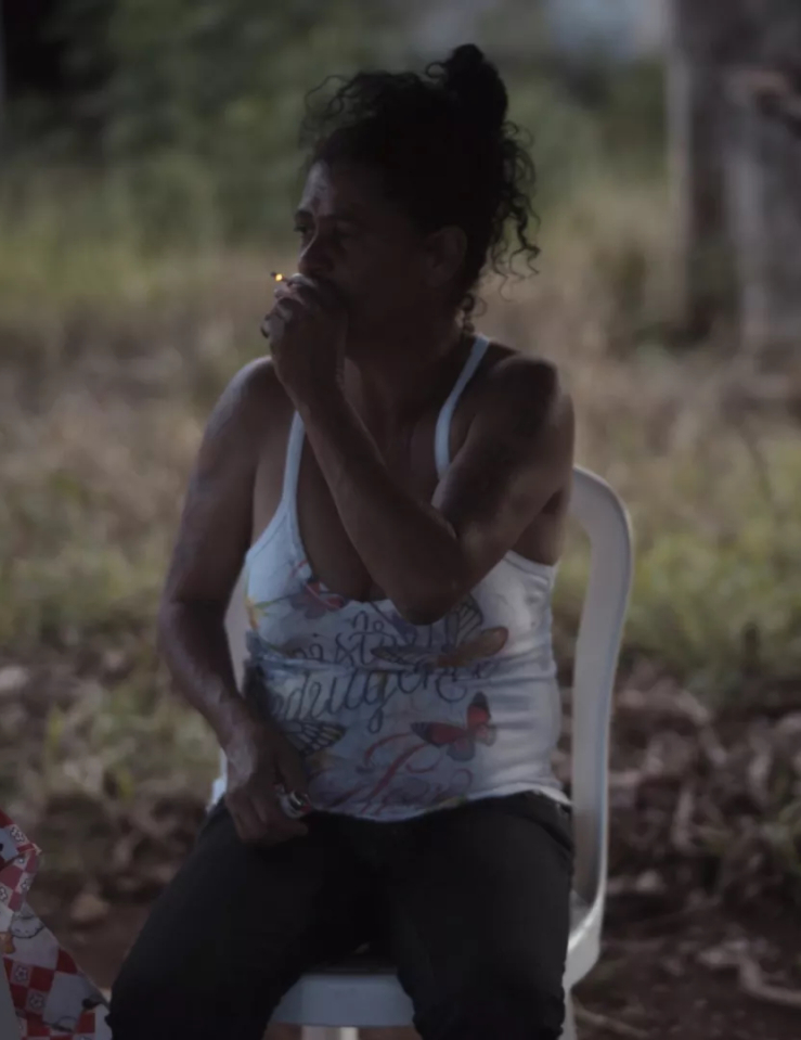A dark skinned woman with dark curly hair in a messy bun wearing jeans and a ratty tank top sits on a plastic chair outside holds a lit cigarette up to her lips